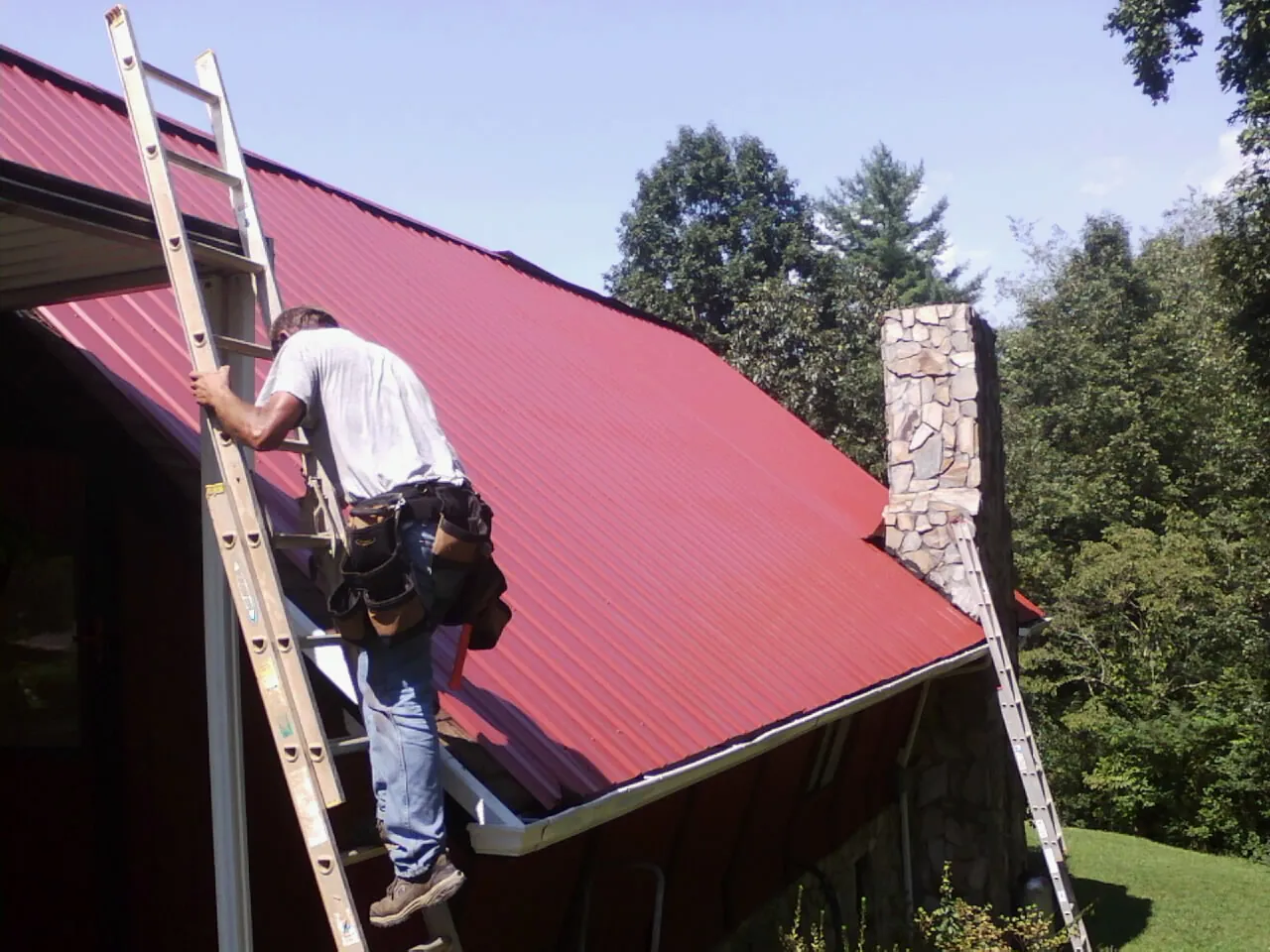 Roofer on a ladder beside a red metal roof and stone chimney — Soffit & Fascia Repair by Roofing Brothers in Lusby